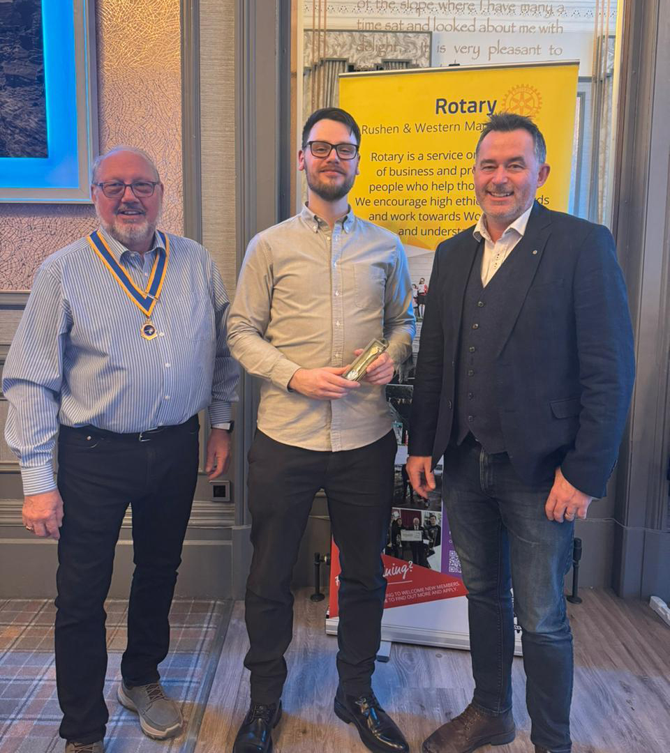 Photograph of Mike Johnson, Darren Barklie and Matt Williams standing together in a row in front of a yellow Rotary Club sign. Mike is wearing his yellow and blue Rotary Club presidential medal, and Darren is holding a spoon that has been presented to him.
